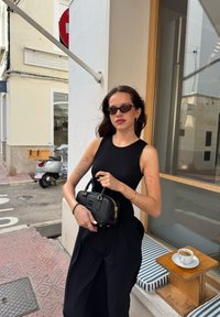 Czarny prążkowany top bez rękawów i długa czarna spódnica, trzymając małą czarną torebkę z złotymi akcentami. Na drewnianym stole obok stoi biała filiżanka espresso.
