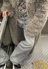 Manteau gris poilu avec manches surdimensionnées, haut étincelant en sequins argentés en dessous, et pantalon gris à jambes larges, complété par des chaussures noires.