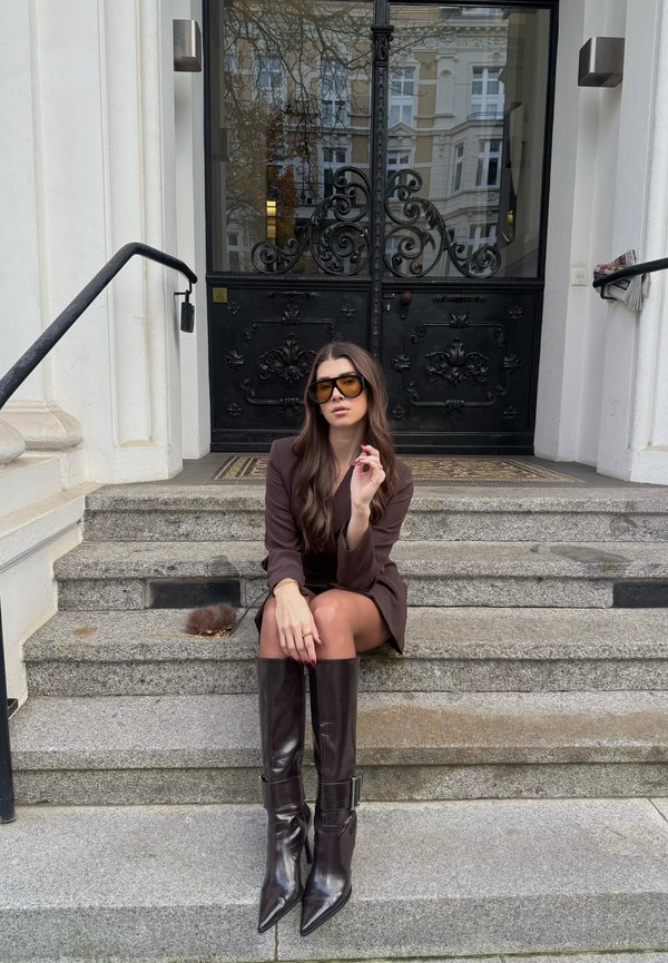 Brown blazer dress paired with knee-high, glossy dark brown heeled boots. The setting features stone steps and an ornate black door.