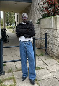 Casaco oversized preto com bordas desfiadas, camisa branca, jeans azul de perna larga e sapatos pretos. Gorro cinza com padrão e óculos azuis.