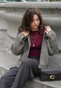 Sivý sako s vrkočovým vzorom so zipsom vpredu a veľkými vreckami, nosené cez bordový top. Tmavosivé nohavice s širokými nohavicami a čierna kabelka.