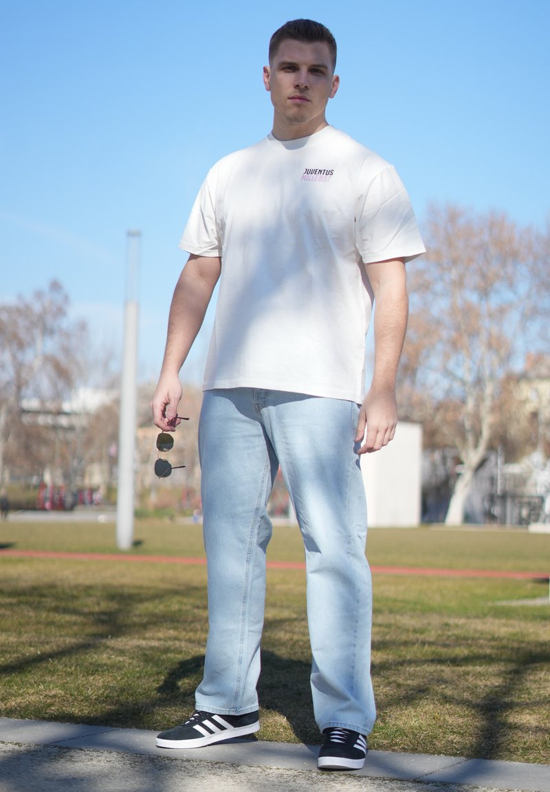 Jeune homme debout en plein air portant un t-shirt blanc Juventus, un jean bleu clair, des baskets noires Adidas, tenant des lunettes de soleil dans sa main droite.