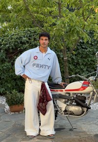 Jaqueta oversized azul claro com o texto "FENTY", calças largas brancas, bandana vermelha e sandálias azuis, posando ao lado de uma motocicleta. Folhagem verde ao fundo.
