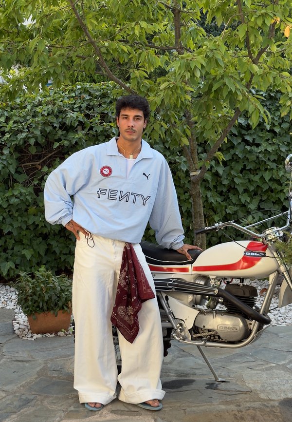 Lyseblå oversized jakke med "FENTY" tekst, hvide brede bukser, rød bandana og blå sandaler, poseret ved siden af en motorcykel. Grøn beplantning i baggrunden.