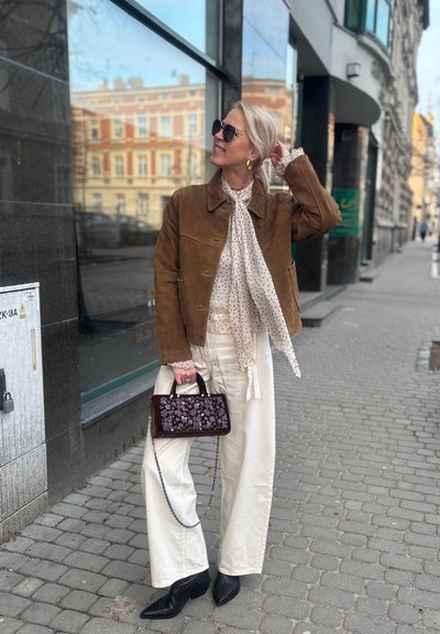 Mujer con chaqueta marrón, pañuelo de lunares, pantalones blancos, botas negras y gafas de sol sosteniendo un bolso floral en la acera de la ciudad.