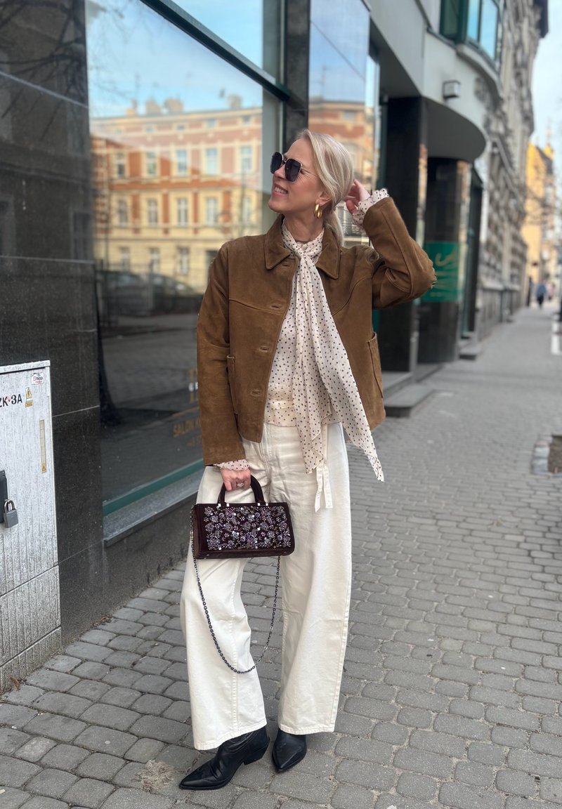 Femme en veste marron, écharpe à pois, pantalon blanc, bottes noires et lunettes de soleil tenant un sac à main fleuri sur un trottoir en ville.