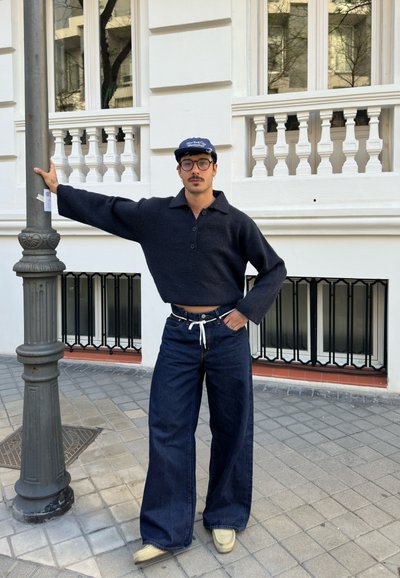 Suéter corto de punto negro con cuello y botones, combinado con jeans de mezclilla oscuros de pierna ancha. Accesorios en azul marino y zapatos claros.