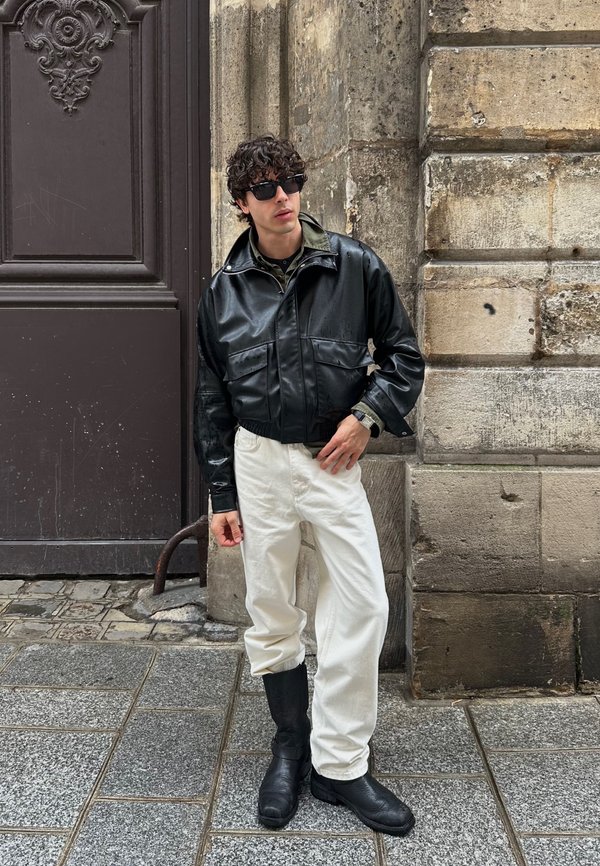 Junger Mann mit lockigem Haar, der eine schwarze Lederjacke, weiße Hose, schwarze Stiefel und Sonnenbrille trägt, steht an einer Steinmauer in einer Stadtstraße.