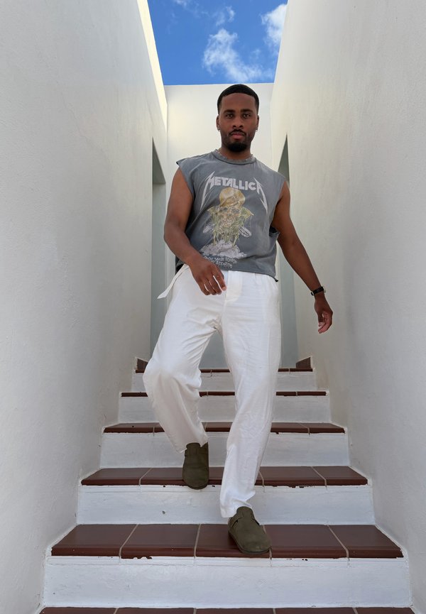 Uomo che scende una scala con piastrelle bianche tra pareti bianche, indossa una maglietta senza maniche dei Metallica, pantaloni bianchi e scarpe scure sotto un cielo azzurro.