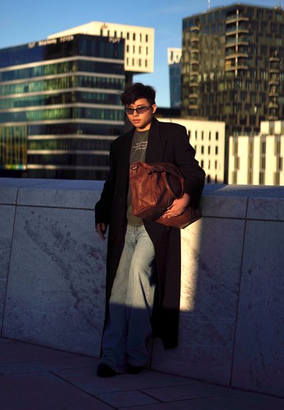 Largo abrigo negro, camiseta oscura, jeans anchos color azul claro y bolso de cuero marrón. Gafas de sol y edificios de fondo.