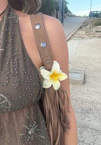 Primer plano de una persona que lleva un vestido de lentejuelas verdes y un bolso de hombro con flecos marrón, con una flor de plumeria amarilla y blanca adjunta.