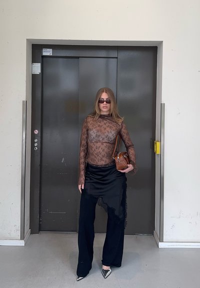 Mujer con blusa marrón floral transparente, falda negra sobre pantalones, zapatos plateados puntiagudos, gafas de sol y sosteniendo un bolso marrón, de pie junto a puertas cerradas del ascensor.