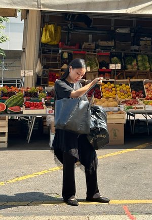 Bolso tote de cuero negro y bolso redondo con logo; mujer con atuendo negro ajustando los bolsos, puesto de mercado con frutas coloridas al fondo.