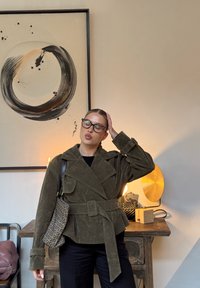 Femme portant des lunettes, vêtue d'une veste ceinturée vert olive et d'un pantalon noir, posant avec la main sur la tête dans une pièce avec une œuvre murale abstraite et une lampe de table.