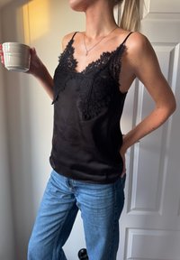 Camisola preta de cetim com renda na gola, alças ajustáveis, combinada com jeans azul claro. Xícara branca segurada na mão esquerda.