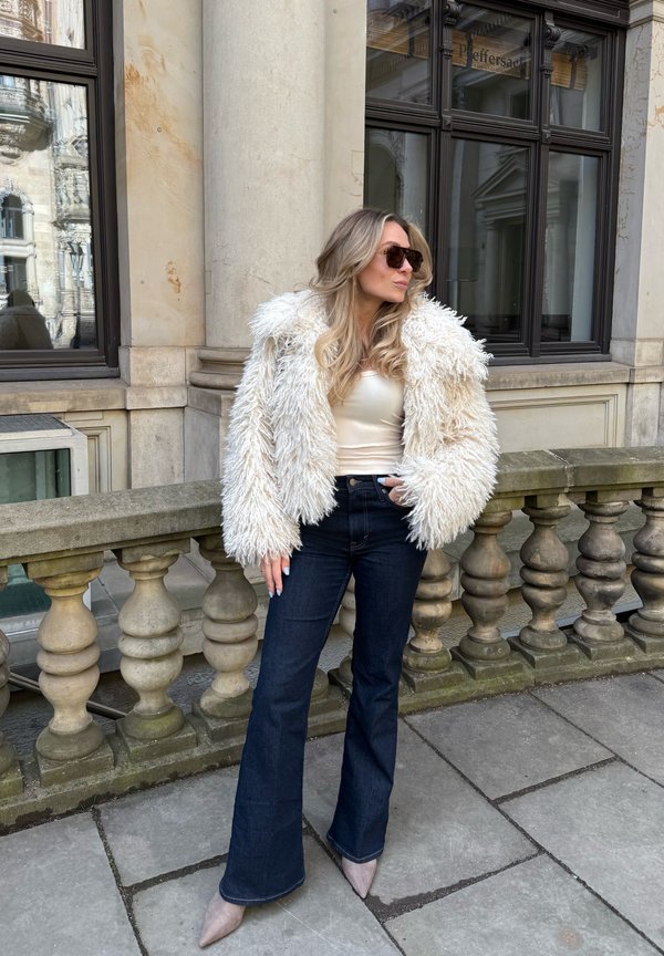 Kvinde iført fluffy hvid jakke, cremefarvet top, mørke vidde jeans og solbriller, står på en stenterrasse med balustrade og store vinduer bagved.