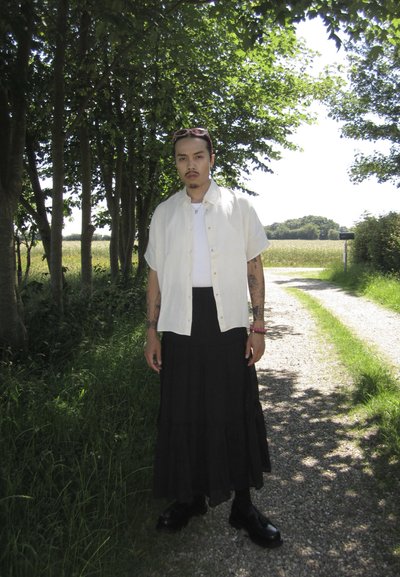 Camisa blanca de manga corta sobre una camiseta blanca ajustada, combinada con una falda larga negra con volantes. Botas negras, con tatuajes visibles en los brazos.