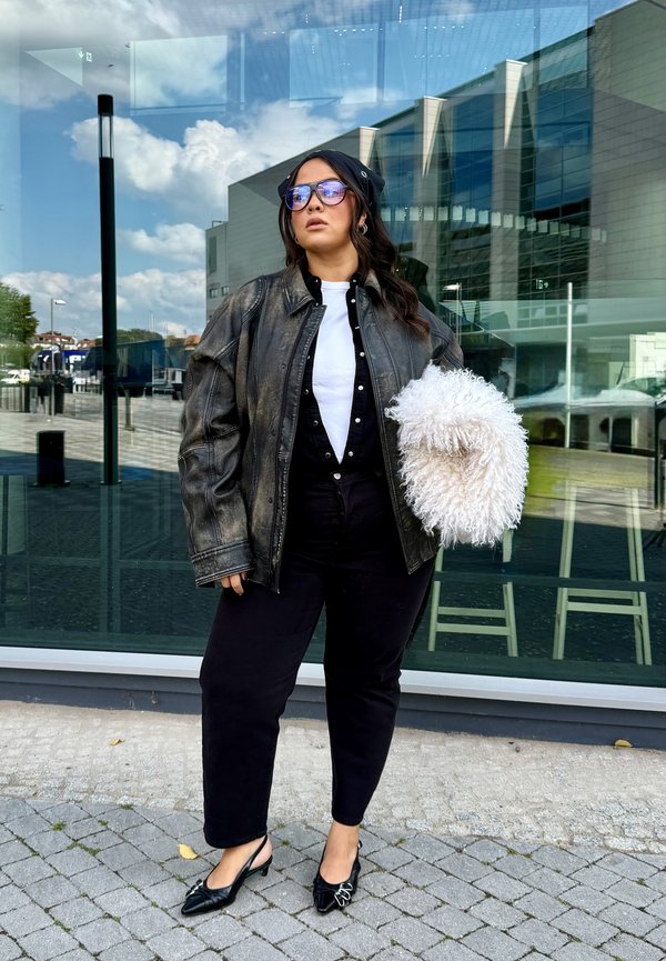 Donna con occhiali da sole, giacca di pelle nera, pantaloni neri e tacchi, che tiene una borsa bianca e pelosa, in piedi su un marciapiede lastricato vicino a un edificio di vetro.