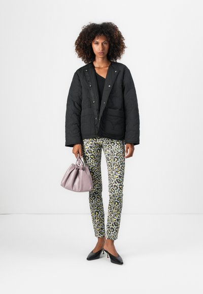 Chaqueta negra acolchada con botones a presión, combinada con pantalones tapered de leopardo en verde y amarillo. Sosteniendo un pequeño bolso lavanda. Tacones negros.