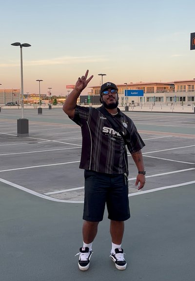 Camisa negra de manga corta con rayas verticales y logo, pantalones cortos negros y zapatillas negras. Persona haciendo un signo de paz, de pie en un área de estacionamiento.