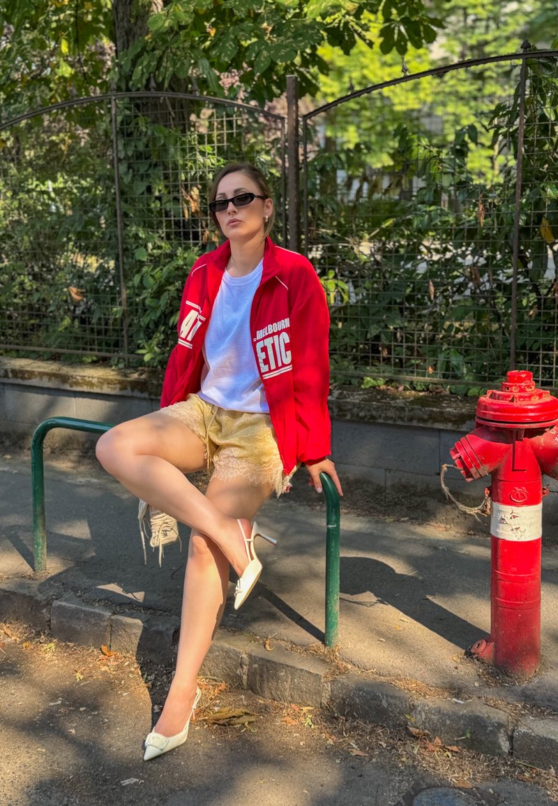 Jeune femme en veste rouge et lunettes de soleil assise sur une rambarde en métal près d'une bouche d'incendie rouge sur un trottoir ombragé avec de la verdure derrière.