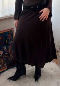 Vestido castanho, fluido, com mangas longas, combinado com um cinto preto. Pés em botas pretas até ao joelho. Tapete de pele branca ao fundo.