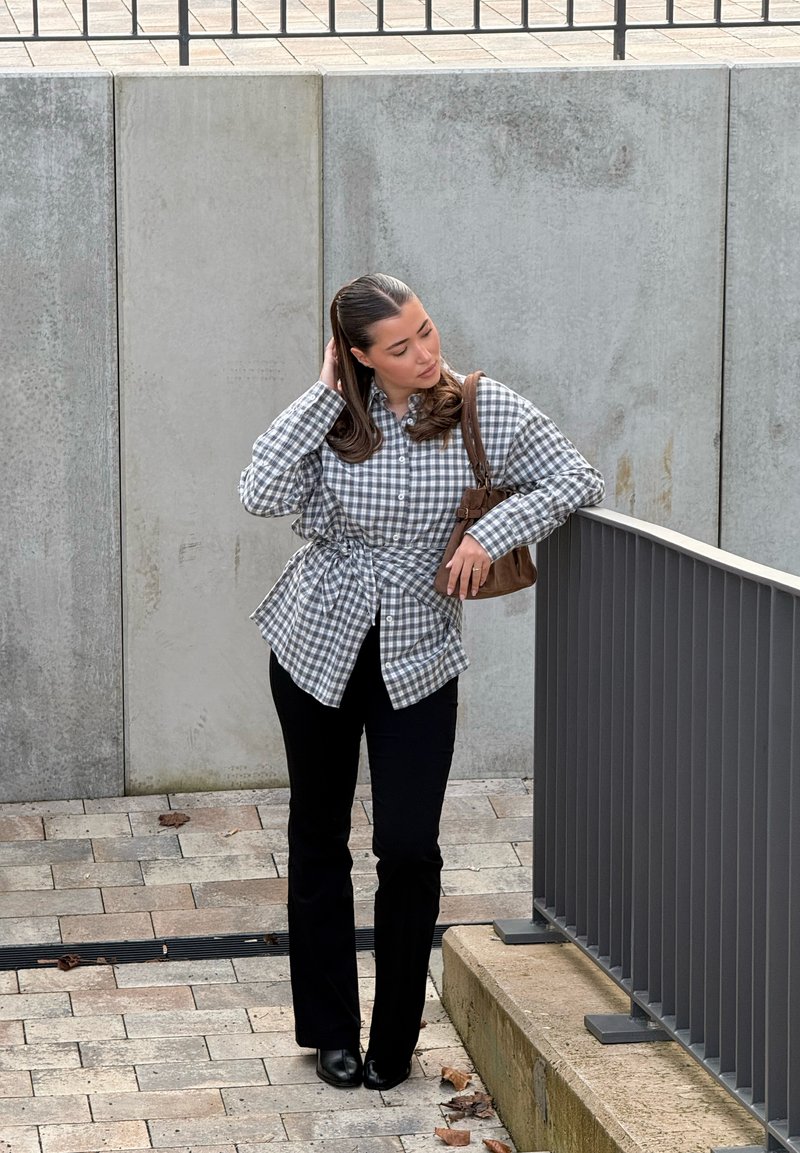 Femme aux cheveux longs portant une chemise à carreaux et un pantalon noir, tenant un sac marron, debout près d'une rambarde métallique grise sur un chemin en pierre.