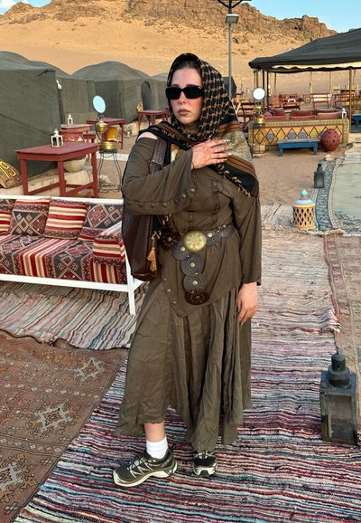 Vestido marrón de manga larga con detalles de botones y cintura ceñida, combinado con zapatillas deportivas y una bufanda estampada en negro y dorado.