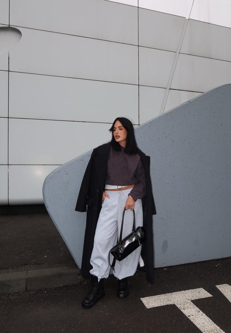 Femme portant un manteau foncé, un pull court, un pantalon de survêtement blanc, des bottes noires, tenant un sac à main noir, debout devant la façade moderne grise d'un bâtiment.