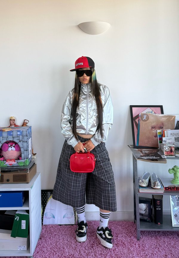 Personne portant une veste argentée, un pantalon large à carreaux, des chaussettes rayées, des baskets noires, une casquette rouge, des lunettes de soleil, tenant un petit sac rouge à l'intérieur.