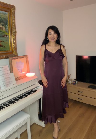 Mujer con un vestido de satén morado, de pie junto a un piano blanco con partituras, una lámpara brillante con forma de cuenco y obras de arte enmarcadas en una sala de estar.