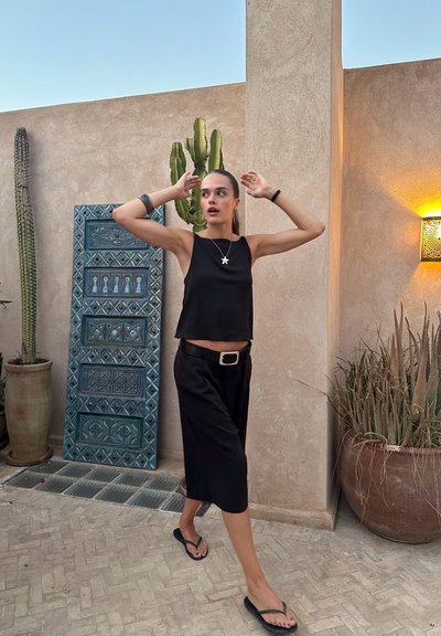 Top negro sin mangas y pantalones cortos negros, combinados con chanclas. Accesorios plateados que incluyen un cinturón y un collar de estrella. El fondo texturizado presenta plantas y una pared decorativa.