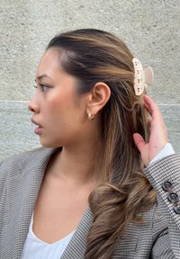 Beige hiusklipsi, jossa on kukkakuvio, pitelee laineilevia hiuksia toiselta puolelta. Ruskea ruudullinen bleiseri ja valkoinen toppi, esillä harmaan taustan edessä.