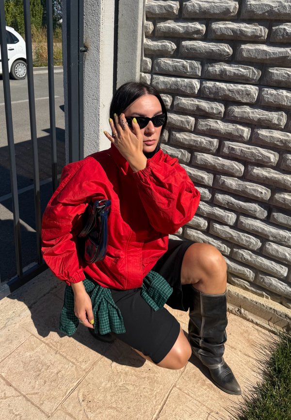Mujer se arrodilla cerca de un muro de piedra con chaqueta roja, falda negra y botas negras, lleva gafas de sol y se toca la cara con una mano.