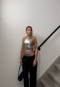 Jeune femme portant un haut argenté dos nu et un pantalon noir, debout près d'un escalier gris, tenant un sac à main noir, regardant sur le côté contre un mur blanc.