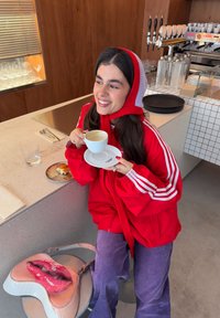 Νεαρή γυναίκα με κόκκινο μπουφάν και κουκούλα που κρατά ένα φλιτζάνι καφέ με πιάτο, καθισμένη σε πάγκο καφέ με ένα γλυκό και μία τσάντα με σχέδιο χείλη κοντά της.