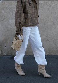 Giacca oversize marrone a quadri, pantaloni bianchi a gamba larga, stivali beige con tacco e una borsa beige strutturata con un design traforato.