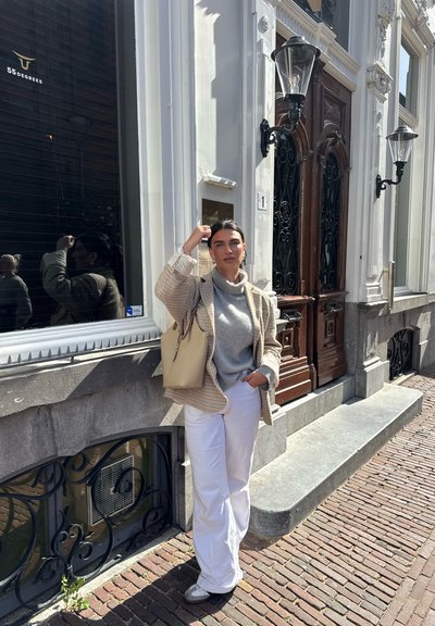 Blazer de cuadros claros sobre un suéter gris, combinado con pantalones blancos. Bolso beige, colocado en una acera cerca de una fachada de edificio decorativa.