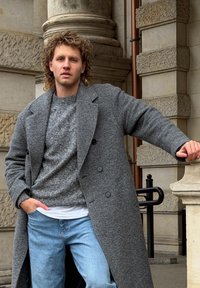 Manteau en laine gris à double boutonnage, porté sur un pull gris texturé et une chemise blanche, associé à un jean bleu clair.