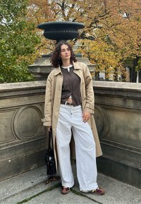 Μπεζ παλτό πάνω από καφέ κάρντιγκαν, λευκά παντελόνια με φαρδύ κόψιμο και καφέ αθλητικά. Μαύρη τσάντα και σκουλαρίκια κρίκοι. Φθινοπωρινά φύλλα στο φόντο.