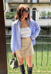 Ανοιχτό μπλε πουκάμισο με μακριά μανίκια πάνω από ένα λευκό crop top, μπεζ φούστα με τσέπες, μαύρες μπότες μέχρι το γόνατο και μια πράσινη τσάντα. Το φόντο περιλαμβάνει νερό.