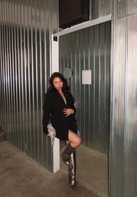 Svart blazer-kjole med dyp V-hals, kombinert med metalliske knelengde støvler. Holder et glitrende tilbehør, satt mot en sølvfarget panelbakgrunn.