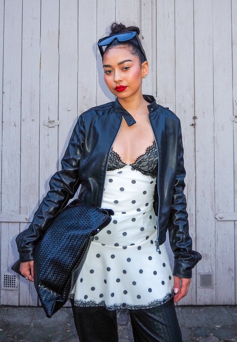 Jeune femme portant un rouge à lèvres rouge, une veste en cuir noire, une robe blanche à pois avec une bordure en dentelle, tenant une pochette noire tissée, debout près d'une porte en bois.