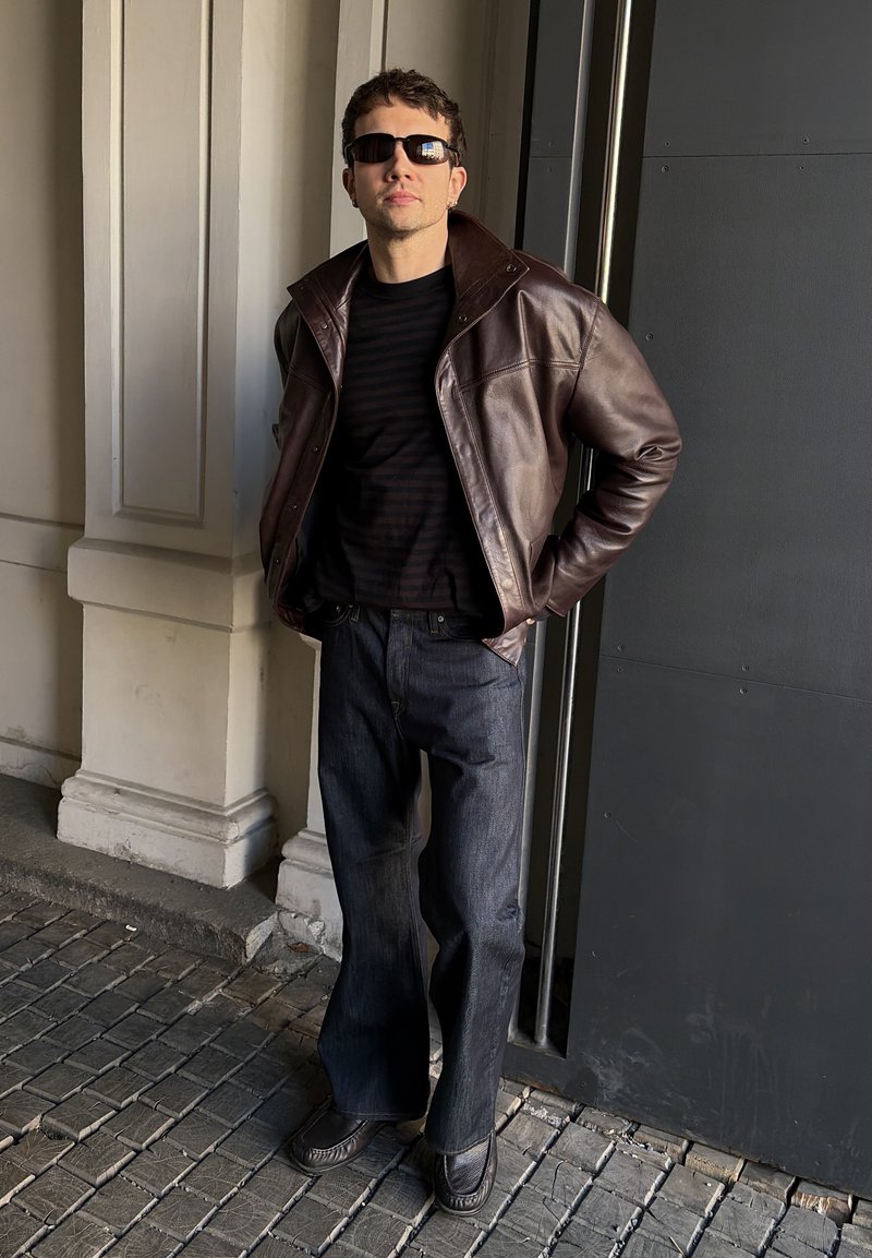Homme portant une veste en cuir marron, une chemise noire, un jean sombre et des lunettes de soleil, debout sur un pavement en pierre près des colonnes d’un bâtiment et d’un mur sombre.