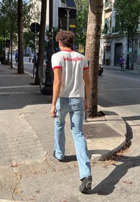 T-shirt bianca con dettagli rossi, con il logo "Wrangler" sulla parte posteriore. Jeans di un leggero blu, che tiene occhiali da sole, mentre cammina su un marciapiede cittadino.