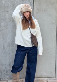 Túlméretezett fehér gyapjúpulóver, sötétkék bő szárú farmer, barna oldaltáska és bézs bolyhos sapka. Simogató anyagok, hétköznapi dizájn.