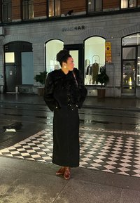 Μαύρο μπουφάν από ψεύτικη γούνα πάνω από μια μακριά μαύρη φούστα, καφέ μπότες, μεγάλα σκουλαρίκια, στέκεται σε ένα διακοσμημένο πεζοδρόμιο κοντά σε ένα κατάστημα τη νύχτα.