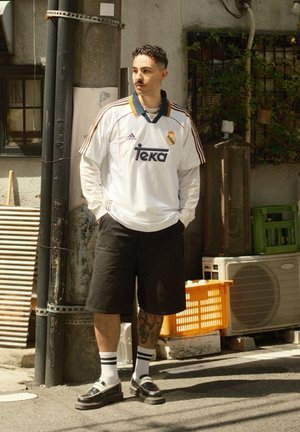 Camiseta blanca del Real Madrid con detalles en azul y amarillo, pantalones cortos negros y zapatos negros con suelas blancas. Medias a rayas y tatuajes visibles.