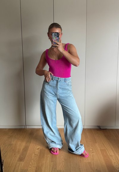 Top de punto fucsia, jeans de pierna ancha azul claro y chanclas rosas. El conjunto se complementa con collares en capas y una funda de teléfono colorida.