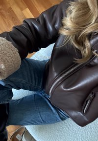 Άτομο που φοράει σκούρο καφέ δερμάτινο μπουφάν με μανσέτες από shearling και μπλε τζιν, κ αθισμένο σε ανοιχτόχρωμη επιφάνεια με υφή.